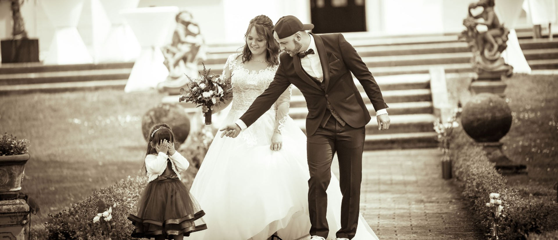 Ein Hochzeitspaar und ein kleines Kind; das Kind verdeckt das Gesicht, alle lächeln, in Sepia.