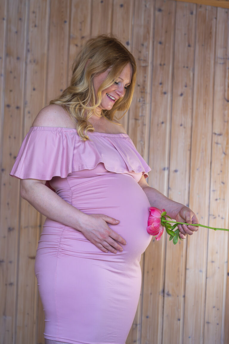 Schwangere Frau lächelnd in rosa Kleid mit Blume, vor holzverkleideter Wand.